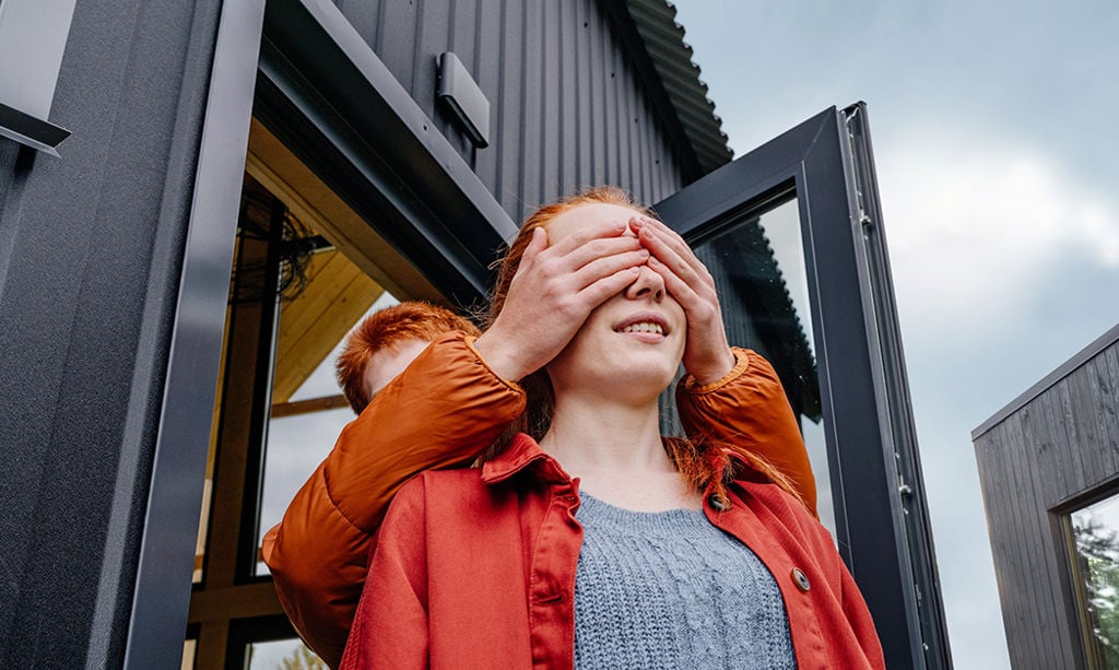 Two people stand in front of the house and the one behind covers the other's eyes with his hands.