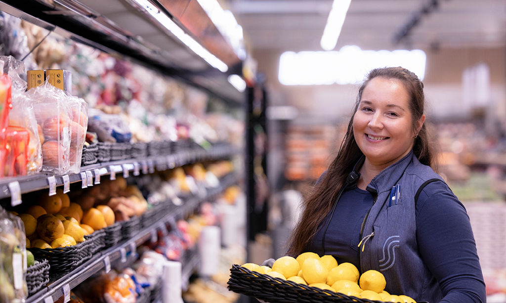 A woman looks at the camera in the refrigerated section of the grocery store. He is smiling and has a fruit basket on his lap.