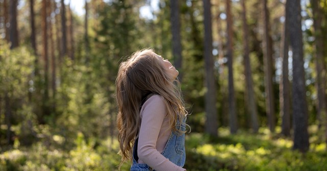 A child with long hair and wearing denim overalls is looking up at the sky in the forest.