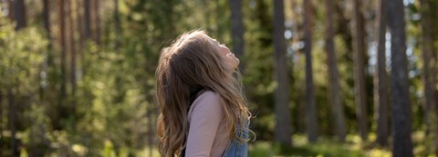  child with long hair and wearing denim overalls looks up to the sky in a summer forest.
