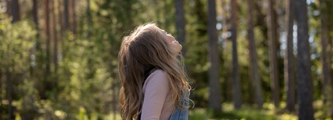 Lapsi, jolla on pitkät hiukset ja yllään farkkuhaalarit, katsoo kesäisessä metsässä taivasta kohti.
