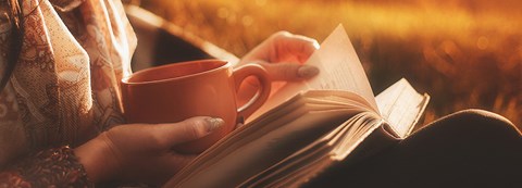 A woman holds a cup of coffee and a book in the autumn sunshine.