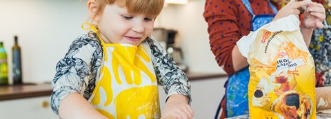 Two children are baking buns. The child in the foreground is rolling the dough on a table with a lot of flour.