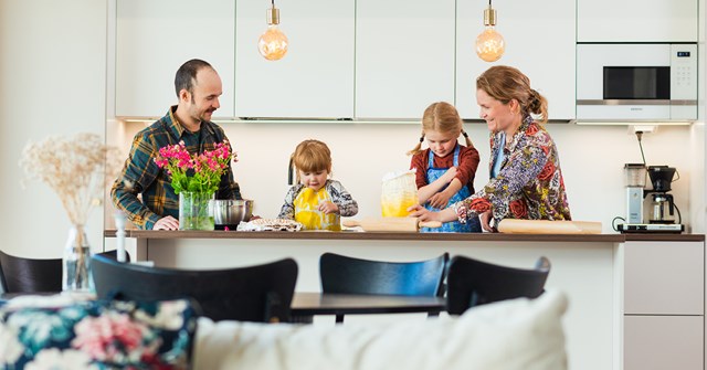 A family of four baking together in the kitchen of their home. There are flowers in a vase, a bag of flour and other baking supplies on the table.