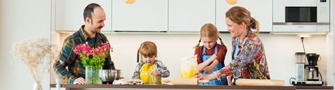 Isä, äiti ja kaksi lasta leipomassa yhdessä kotinsa keittiössä. Pöydällä on kukkia maljakossa, jauhopussi sekä muita leivontatarvikkeita.