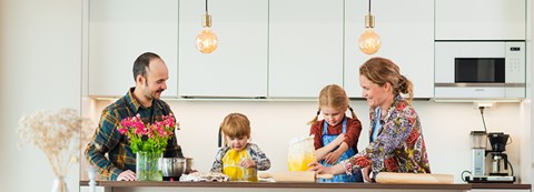 Isä, äiti ja kaksi lasta leipomassa yhdessä kotinsa keittiössä. Pöydällä on kukkia maljakossa ja leivontatarvikkeita. 
