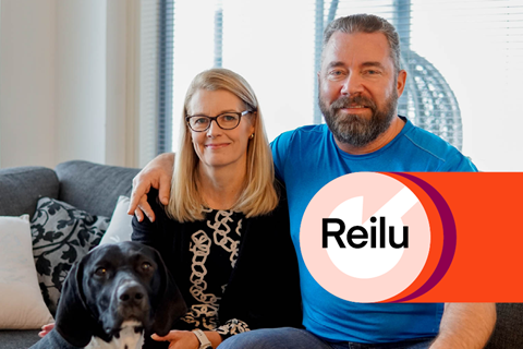 Pasi ja Minna Alajoki istuvat sohvalla koiransa kanssa. Kuvan päällä on Reilu-kaukolämmön punainen elementti.