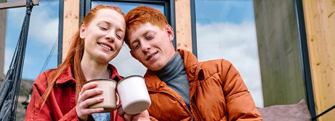 Nuori punatukkainen nainen ja nuori punatukkainen mies kippistävät kahvimukeillaan ja istuvat vierekkäin portailla.