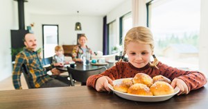 The girl is holding the bun plate that is on the table and looking at the buns. Mother, little sister and father are sitting in the background by the dining table.
