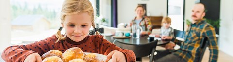 The girl is holding the bun plate that is on the table and looking at the buns. Mother, little sister and father are sitting in the background by the dining table.