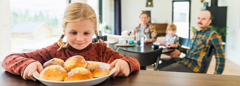 Tyttö pitää kiinni pullalautasesta, joka on pöydällä ja katsoo pullia. Äiti, pikkusisko ja isä istuvat taustalla ruokapöydän ääressä.