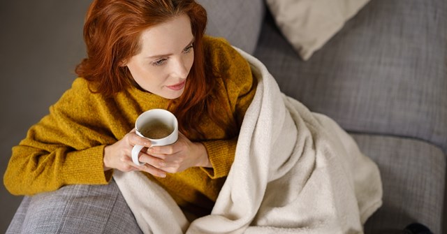 A red-haired woman is sitting on a couch with a blanket and holding a cup in her hands.