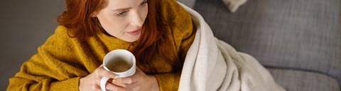 A red-haired woman is holding a white cup and sitting on a couch with a white blanket.