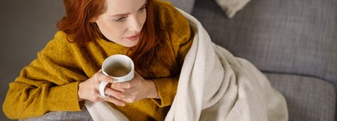 A red-haired woman is sitting on a couch under a blanket and holding a teacup in her hands.