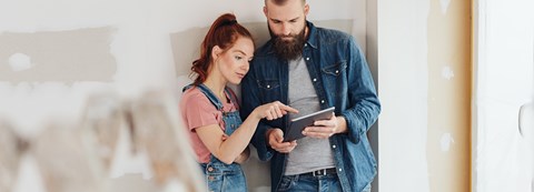 In a room that is currently under renovation, a couple is looking a tablet which the man is holding up.