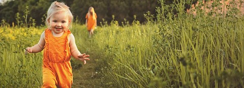 A child runs on a green field. An adult is walking behind.