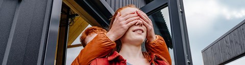 The person behind covers the eyes of the red-haired person standing in front with their hands. The person standing in front is smiling.