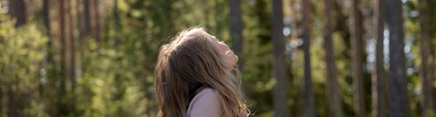 A child with long hair and wearing denim overalls is looking up at the sky in a sunny forest.