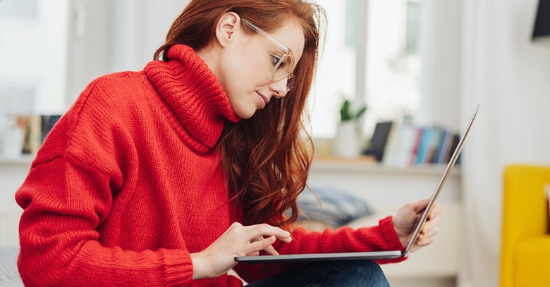A red-haired woman, who is wearing a red sweater is using a laptop which she is holding in her lap.