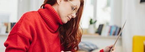 Young red-haired woman, wearing a red sweater, is scrolling through a laptop.