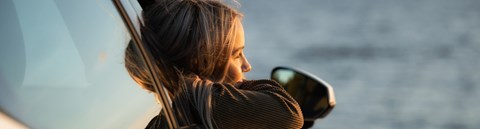 Blond girl is sitting in a car with an open window. The girl is looking to the sea.