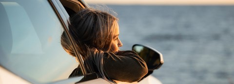 A girl is sitting in a car and looking to the sea. The car's window is open and the girl is leaning outside of it.