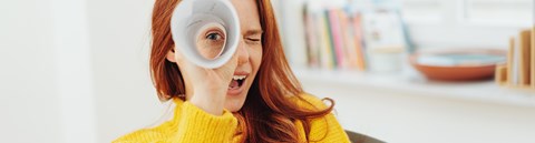 A red-haired woman is looking to the camera through a paper roll. She is wearing a yellow sweater.