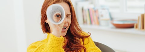 A red-haired woman is sitting on a chair and looking to camera through a paper roll. She is wearing a yellow shirt.