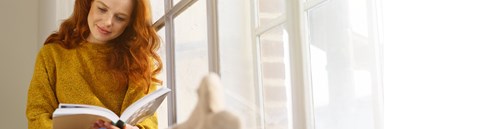 A red-haired woman is sitting in a windowsill and reading a book. She is wearing a yellow sweater.