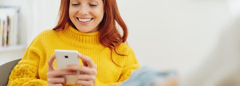 A smiling woman is scrolling through her phone, which she is holding in her hands.