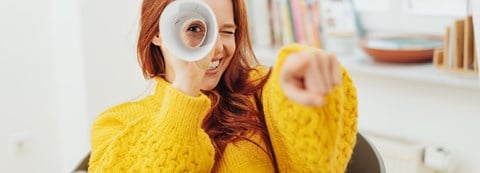 Young red-haired woman is looking straight to the camera through a roll made out of a papersheet.