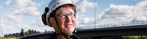 An employee of Oulu Energy is wearing their work gear and stands infront of the Oulu River.