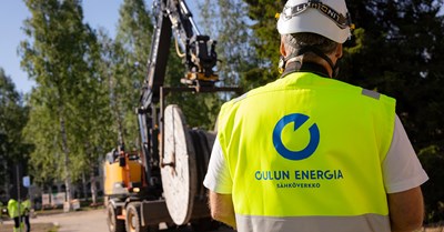 An employee of Oulu Energy is standing and facing away from the camera. There is an excavator in the background.