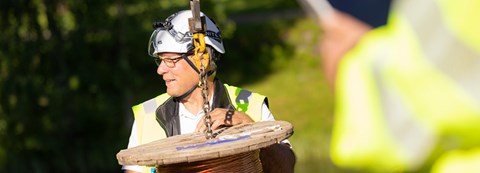 An employee of Oulu Energy is wearing their work gear and there is a cable roll hanging from an iron chain in front of him.