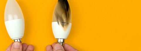 A person is holding two light bulbs in their hands in front of an orange background. The other light bulb has burnt and the other one not.
