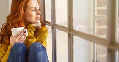 A red-haired woman is holding a coffee mug in her hands and sits on the windowsill and looks out.