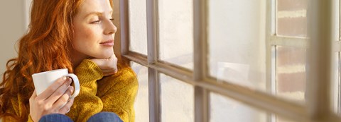 Punatukkainen nainen istuu kahvikuppi kädessään ikkunalaudalla ja katselee ulos.