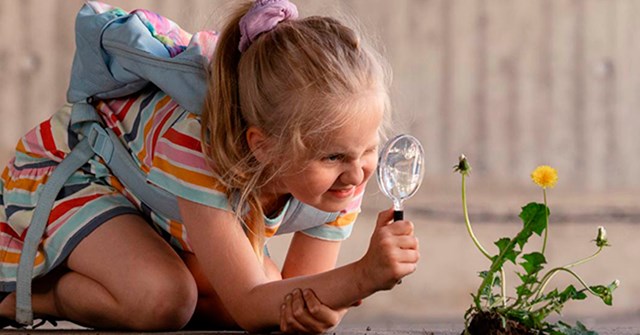 A girl examines a yellow dandelion with a magnifying glass.