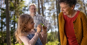 A child and grandparents look at a leaf that has fallen from a tree in the forest and smile.