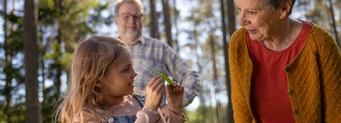 Lapsi ja isovanhemmat katsovat puusta tippunutta lehteä kesäisessä metsässä.