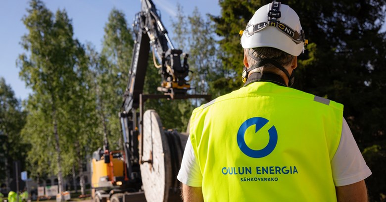 In the photo, a man is standing wearing a safety helmet and wearing a safety vest. The man looks at the excavator in the background.