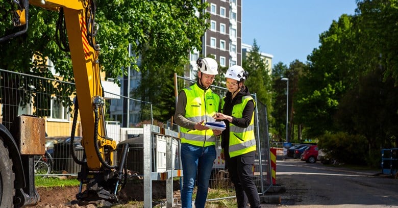 Oulun Energian kaukolämmön työmaalla kaksi työntekijää katsoo papereita yhdessä. Taustalla on kaivinkone. 
