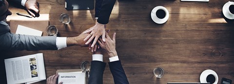 The dark brown conference room table, pictured from above, with hands outstretched together from all sides. The desk also holds a variety of office supplies, including pens, a laptop, a notepad.