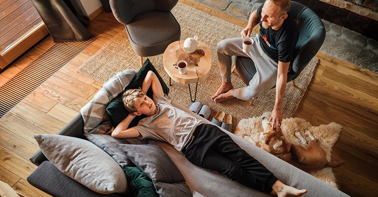  A young child is lying on the couch and looking up. The parent sits next to him in an armchair and laughs.