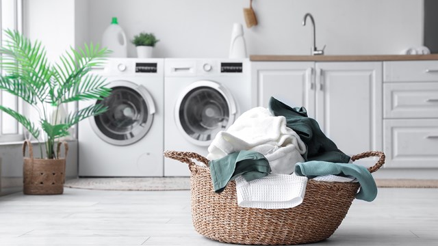 Pyykkikori täynnä puhtaita tekstiilejä modernissa ja valoisassa kodinhoitohuoneessa, jossa pesukone ja kuivausrumpu on sijoitettu vierekkäin.