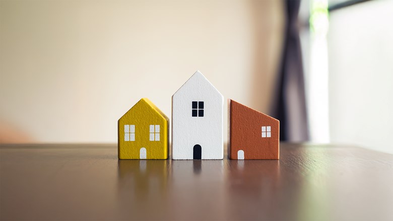 Kolme eriväristä puista pienoismallitaloa rivissä pöydällä, symboloiden erilaisia koteja.