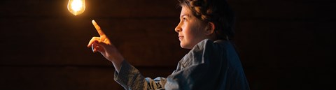 A young girl in pigtails touches a light bulb hanging from the ceiling with her finger.