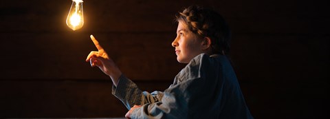 A young girl in pigtails touches a swinging light bulb hanging from the ceiling with her finger.