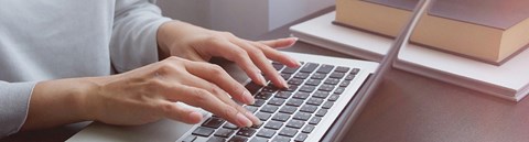 Nainen on kirjoittamassa läppärin näppäimistöllä. Kuva on rajattu niin, että vain osa vartalosta sekä kädet näkyvät.