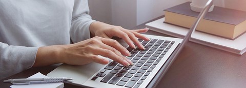 Nainen on tekemässä töitä läppärillä. Kuva on rajattu niin, että vain osa vartalosta sekä kädet näkyvät.